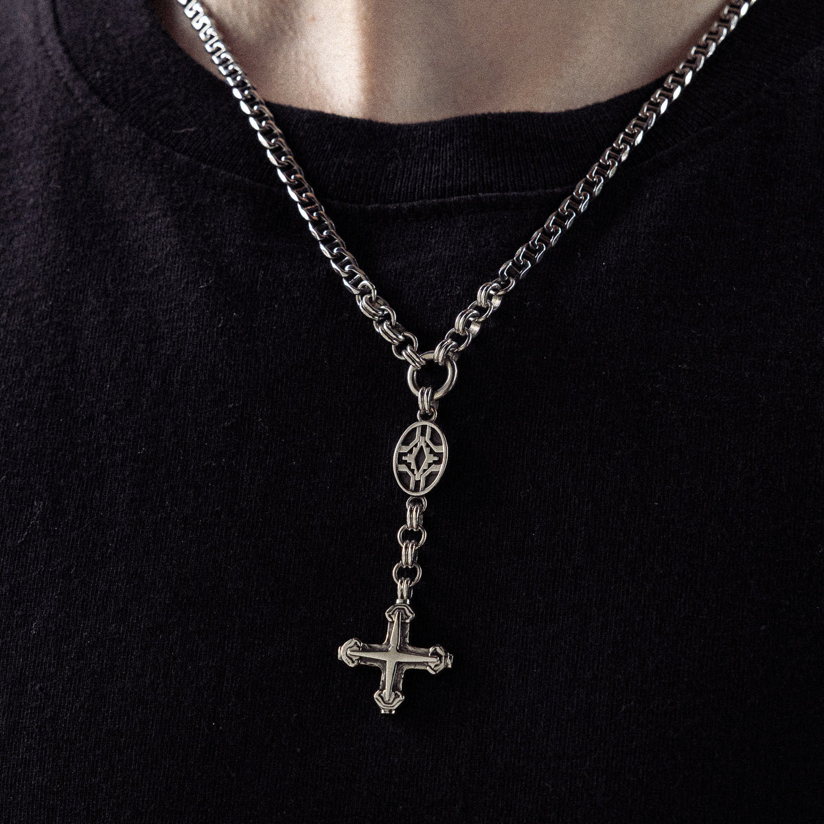 Silver-toned chain necklace with ornamental oval link and dangling cross pendant worn on black crew neck shirt.