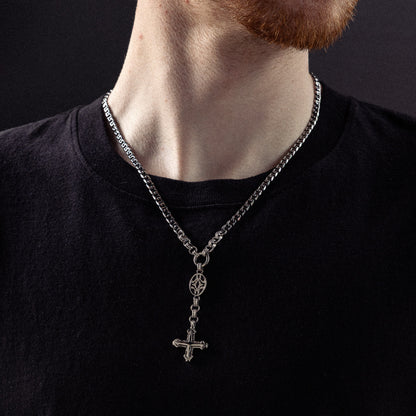 Man wearing silver chain necklace with ornate oval pendant and inverted cross charm on black shirt.
