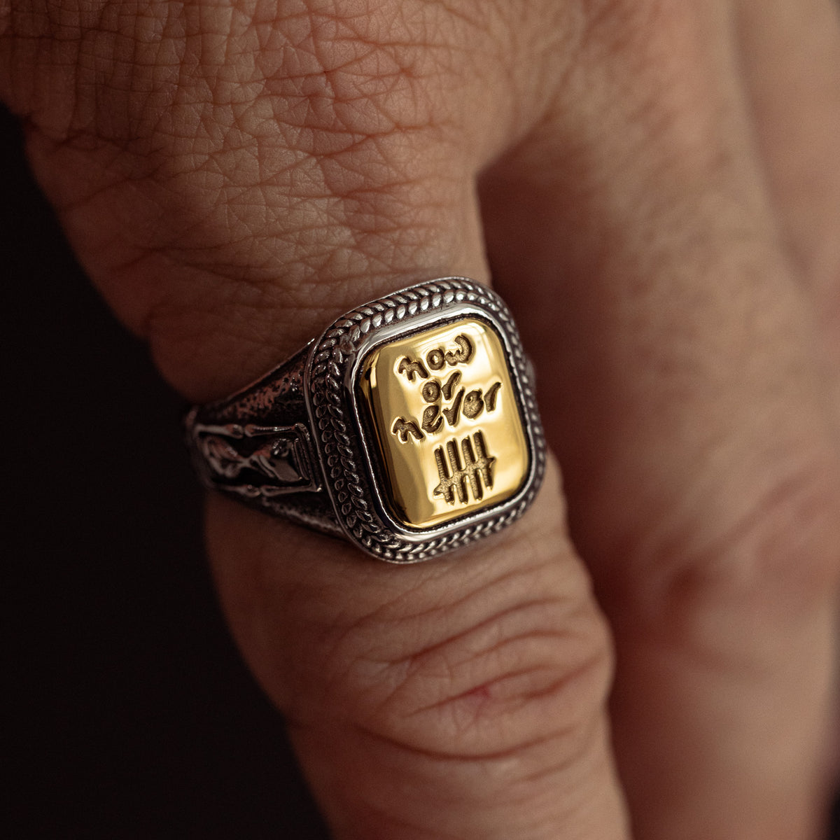 Silver and gold statement ring with "Now or Never" engraved on rectangular face, worn on finger.