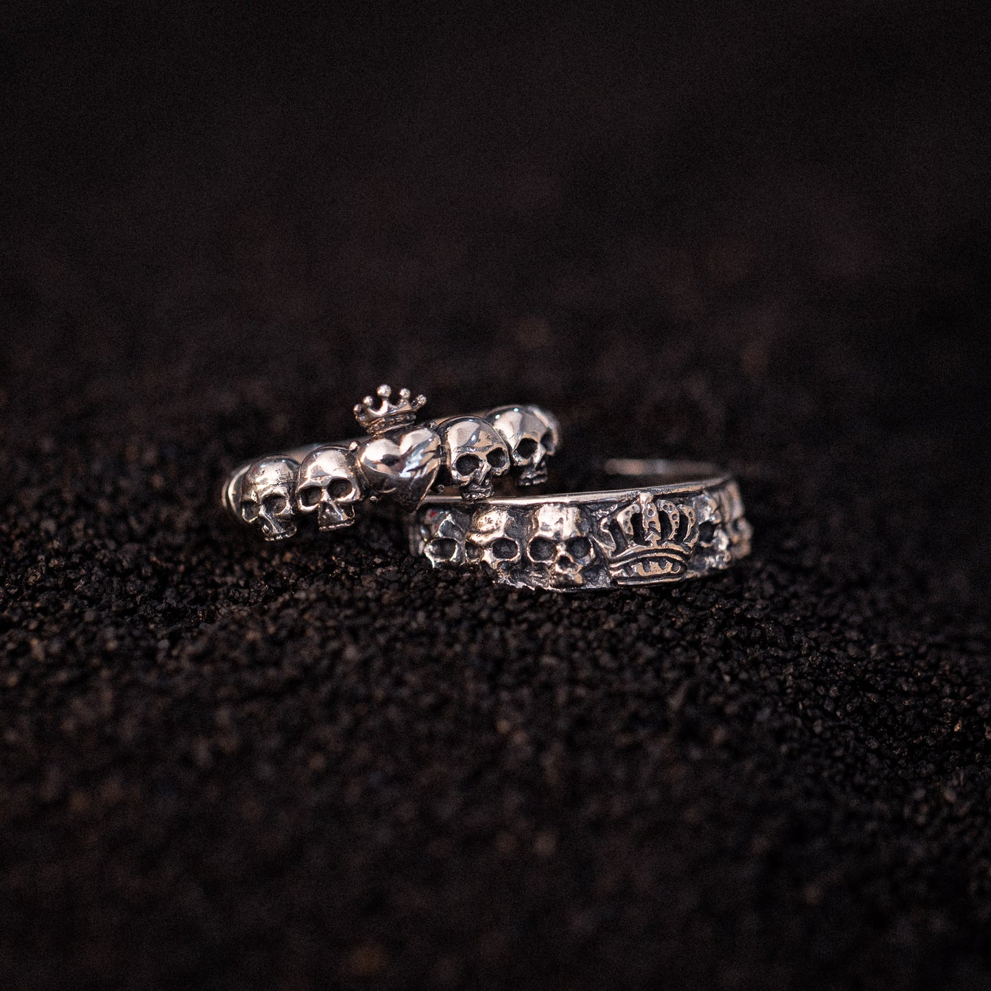 Two silver rings with skull designs and crown details on dark textured surface.