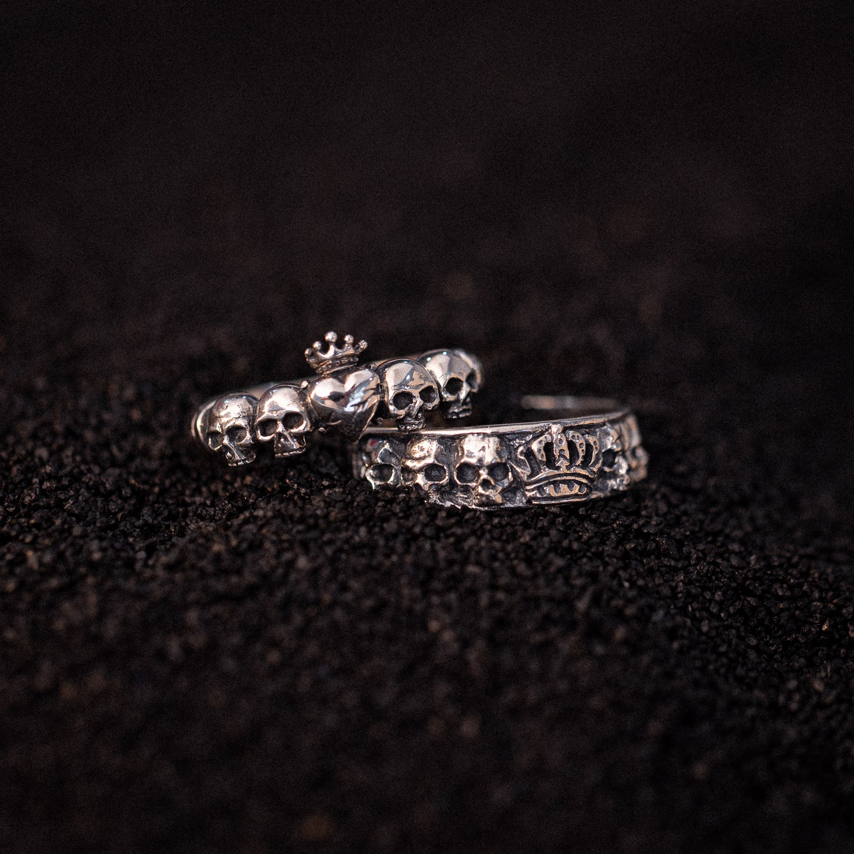 Two silver rings with skull designs and crown details on dark textured surface.