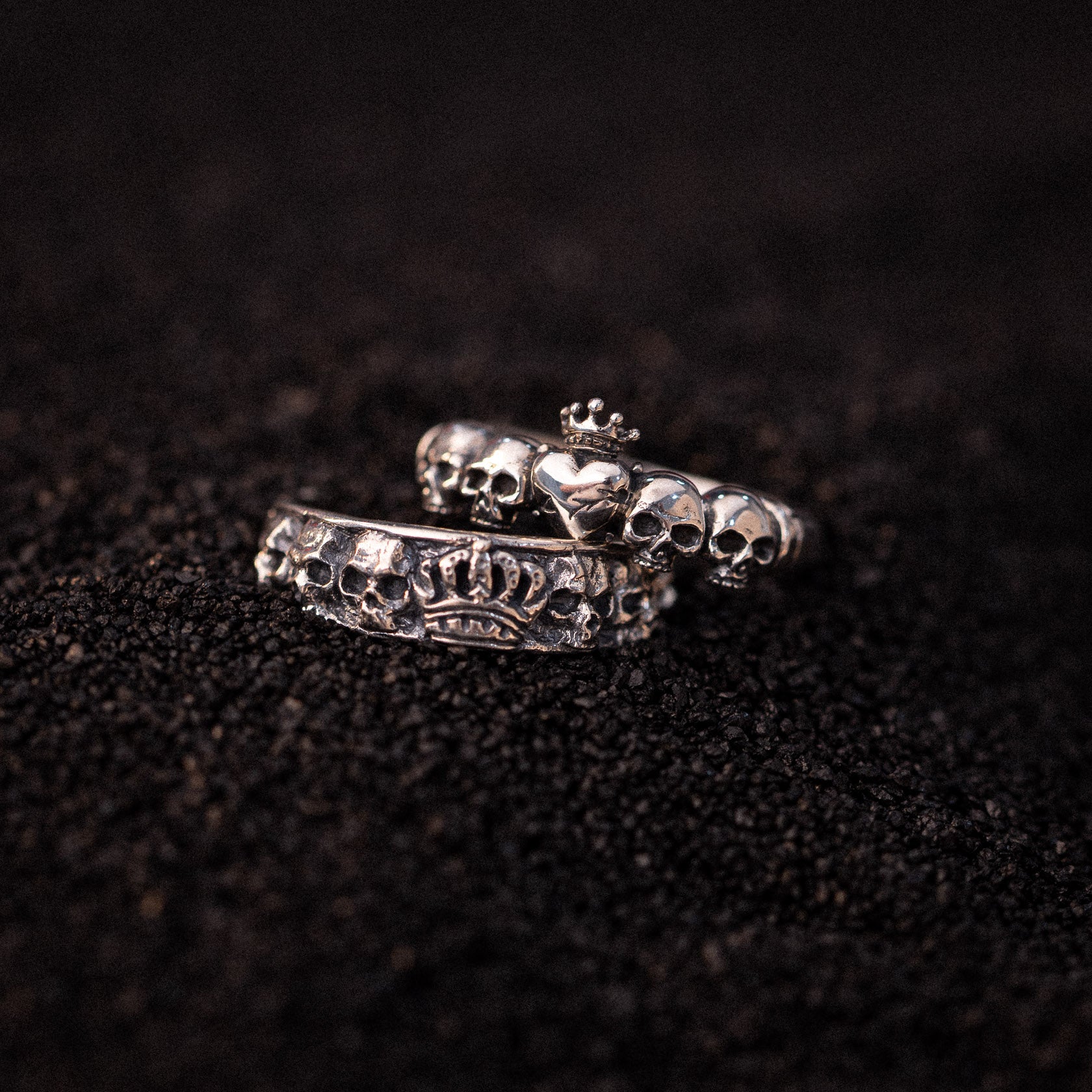 Two silver rings with skull and heart motifs displayed on dark textured surface.