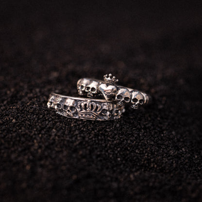 Two silver rings with skull and heart motifs displayed on dark textured surface.